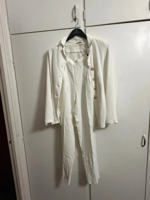 Vit kostym med kavaj och byxor  från PRISMA - Säljer en stilren vit kostym med matchande kavaj och byxor. Kavajen har klassisk passform, knappar framtill och långa ärmar. Byxorna är raka och höga i midjan. Perfekt för dig som vill ha en clean och fräsch look.