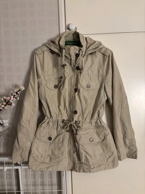 Isolde jacka - En väldigt fin jacka från Isolde med många detaljer. En knapp har försvunnit (kolla bild) men det finns extra knappar att sy på. Den är justerbar i midjan och luvan. Den har dragkedja + knappar. Färgen är typ en blandning mellan beige och khaki vetinte vad den kallas för. Storlek 42! Pris går att diskutera.