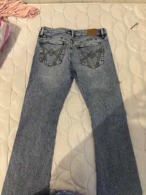 Ljusblå bootcut jeans med broderi - Säljer ett par ljusblå bootcut jeans med snygga broderade detaljer på bakfickorna. Jeansen har klassisk femficksdesign och är tillverkade i denim med lätt tvättad look. Perfekta för dig som gillar en avslappnad men trendig stil.