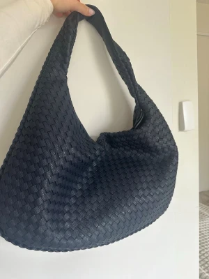 Marinblå flätad axelväska i läderstil - Snygg svart axelväska med flätad design och mjuk, rundad form. Väskan har bred axelrem och är tillverkad i material som liknar läder. Perfekt för dig som gillar trendiga och stilrena accessoarer.