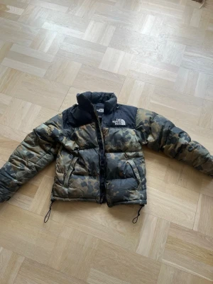Camo pufferjacka från The North Face - Säljer en fet pufferjacka från The North Face med camomönster i grönt, brunt och svart. Jackan har svarta axelpartier, hög krage och dragkedja framtill. Perfekt för kalla vinterdagar och riktigt street.