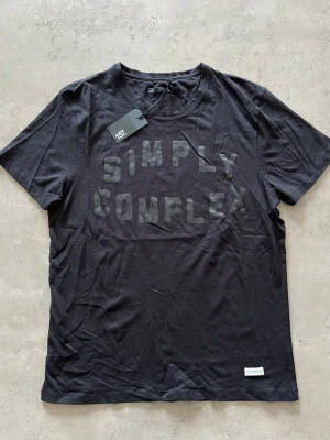 Svart t-shirt från 157 med tryck - L - Svart t-shirt från 157 med texten 'SIMPLY COMPLEX' i ton-i-ton tryck på bröstet. Klassisk rund halsringning och korta ärmar. Tillverkad i mjuk bomull som känns skön mot huden. Perfekt för en clean och stilren look.