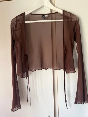 Brun meshkofta från H&M - Säljer en brun transparent meshkofta från H&M med långa ärmar och knytband framtill. Koftan har vågig kant nertill och är perfekt för lager på lager-stil. Tunn och luftig design som ger en trendig look.