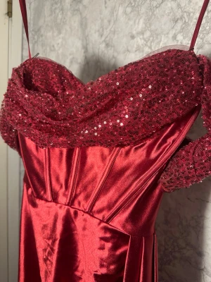 Röd långklänning med paljetter - Elegant röd långklänning med smala axelband och glittrig paljettdekor över bysten. Klänningen har en draperad detalj på sidan och är tillverkad i ett glänsande satinliknande material. Perfekt för dig som vill sticka ut och känna dig extra glamorös.