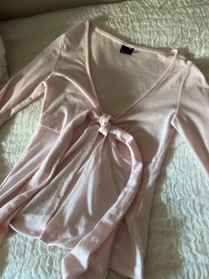 Ljusrosa omlottopp med knyt  - Supersöt ljusrosa omlottopp från Gina med lång ärm och knyt framtill. Tunn och mjuk i materialet, perfekt för sommaren eller över ett linne. Djup v-ringning och figurnära passform ger en feminin vibe.