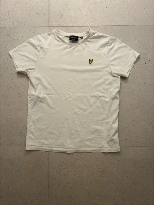 Vit t-shirt från Lyle & Scott - Vit t-shirt från Lyle & Scott med klassisk passform och korta ärmar. T-shirten har en liten gul broderad logga på bröstet och är tillverkad i mjuk bomull. Perfekt för en clean och stilren look.