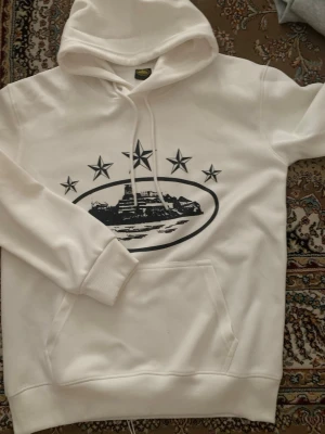 Corteiz Hoodie  - Säljer en vit hoodie med huva och känguruficka. På bröstet finns ett svart tryck med fem stjärnor och ett motiv av en byggnad eller landskap inuti en oval. Dragsko i huvan och ribbade muddar vid ärmslut. Perfekt för dig som gillar streetwear och grafiska prints.