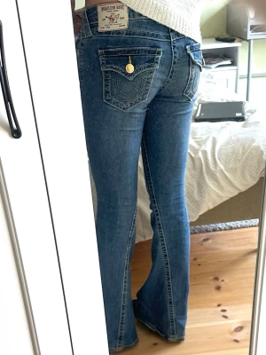 Jeans från True Religion - Säljer ett par blå bootcut jeans från True Religion med ikoniska kontrastsömmar och guldknappar på bakfickorna. Jeansen har låg midja och klassisk femficksdesign. Materialet är denim i bomull med lite stretch för skön passform. Stl 25, sparsamt använda, inget tecken på slitage, som nya. 