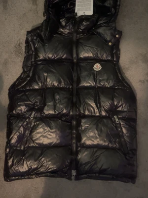 Moncler väst - Snygg svart dunväst från Moncler med glansig finish och klassisk logotyp på bröstet. Västen har två dragkedjeförsedda fickor och hög krage med huva. Perfekt för lager på lager och streetstyle. Skriv gärna vid frågor🙂