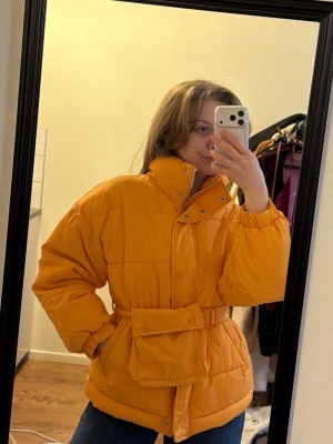 Gul/mustard pufferjacka med bälte - Snygg gul pufferjacka med hög krage och bälte i midjan som ger en schysst siluett. Jackan har stora fickor framtill och stängs med både dragkedja och knappar. Perfekt för kalla dagar när du vill sticka ut lite extra.