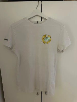 Vit Bajen t-shirt med tryck - Vit t-shirt från Hammarby IF med klubbens emblem tryckt på bröstet och stort grönt BAJEN 12-tryck på ryggen. Klassisk rund hals och korta ärmar. Perfekt för dig som vill visa ditt stöd på matchen eller i vardagen.