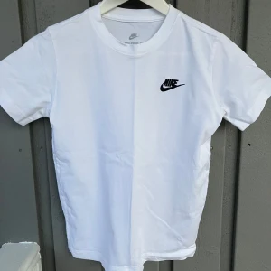 Vit Nike T-shirt med logga - Klassisk vit T-shirt från Nike med svart logga på bröstet. T-shirten har rund halsringning och korta ärmar. Tillverkad i mjuk bomull för en bekväm känsla. Perfekt basplagg för en sportig och stilren look. Barnstorleken motsvarar en tight sittande XXS.