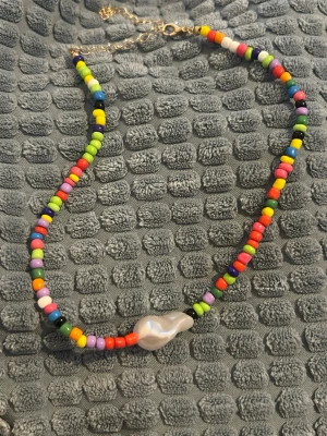 Färgglatt pärlhalsband med barockpärla - halsband med små färgglada plastpärlor i regnbågens alla färger och en stor oregelbunden vit barockpärla i mitten. Halsbandet har guldfärgat justerbart lås och sitter som en choker. Perfekt för dig som vill sticka ut med en lekfull accessoar.