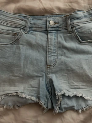 Ljusblå jeansshorts med fransar - Säljer ett par ljusblå jeansshorts med fransiga benslut och slitna detaljer på bakfickorna. Klassisk femficksmodell med knapp och dragkedja framtill. Perfekta för varma dagar och en avslappnad stil. Låg-mitt midjad passform på kroppen. 