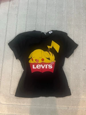 Svart Levi's x Pokémon Pikachu T-shirt - Säljer en svart T-shirt från Levi's x Pokémon med en stor tryckt Pikachu som vilar ovanpå Levi's-loggan i rött och vitt. T-shirten har korta ärmar och är gjord i mjuk bomull. Perfekt för dig som älskar Pokémon och vill ha en unik collab i garderoben.