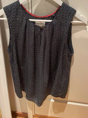 Mönstrad ärmlös topp i strl 40 - Snygg ärmlös topp från Holly & Whyte by Lindex i mörkblått med vitt grafiskt mönster. Toppen har rund halsringning med en röd kantdetalj och är tillverkad i mjuk polyester. Perfekt för en trendig och avslappnad stil.