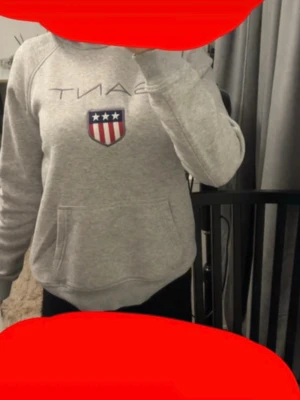Grå hoodie från Gant med logga - Säljer en grå hoodie från Gant med klassisk logga och sköld med stjärnor och ränder på bröstet. Hoodien har en stor magficka och ribbade muddar vid ärmslut och nederkant. Perfekt för en avslappnad och sportig stil.