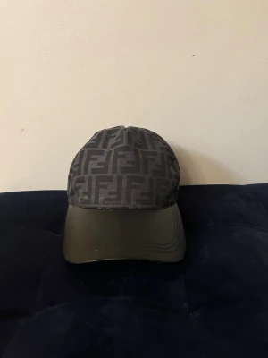 Svart Fendi keps med FF-mönster - Snygg svart keps från Fendi med det klassiska FF-logomönstret över hela kepsen och justerbart band bak i läder. Skärmen är i svart och materialet känns lyxigt och stilrent. Perfekt accessoar för dig som gillar exklusiva detaljer.