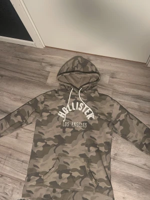 Camouflage hoodie från Hollister - Snygg hoodie från Hollister med grått och beige camouflage-mönster och vit text på bröstet. Klassisk huva med snörning och stor magficka framtill. Perfekt för en chill och avslappnad stil.