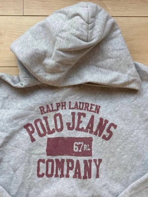 Extremt sällsynt Grå hoodie från Ralph lauren - Snygg grå hoodie från Ralph Lauren med stor vinröd logga och text på bröstet. Klassisk känguruficka framtill och ribbade muddar vid ärmar och nederkant. Perfekt för en avslappnad och sportig stil.