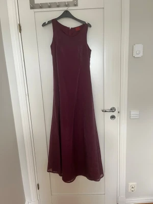 Vinröd långklänning i sidenlook - Elegant vinröd långklänning med ärmlös design och rund halsringning. Klänningen har en lätt glansig yta som ger en exklusiv känsla och ett vackert fall. Jättefin till bal eller annat festligt tillfälle. Jättefint skick! 