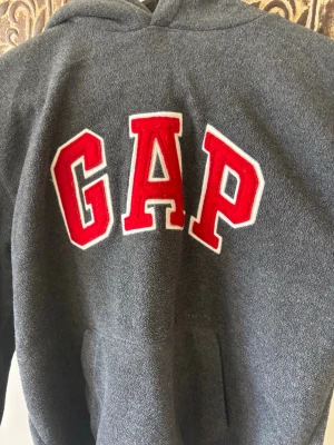 Mörkgrå hoodie från GAP med röd logga - Säljer en mörkgrå hoodie från GAP med stor, röd broderad logga på bröstet. Jättefin men kommer inte till användning. Öppen för att prissänka💕💕💕strl 12-13 år tror jag men passar som en xs-s