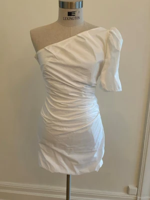 Vit one shoulder klänning Mango - Trendig vit kort klänning med one shoulder-design och puffärm. Klänningen har draperade detaljer och en figurnära passfor. Köpt i Singapore för 150 SGD (1500 SEK). Använd en gång.