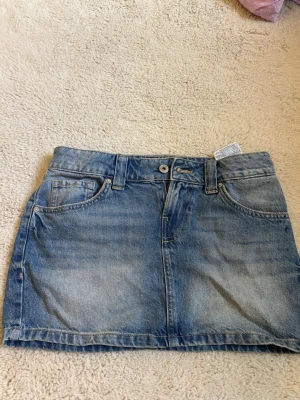 Blå lågmidjad jeanskjol från Bershka - Snygg blå jeanskjol från Bershka med klassisk femficksdesign, bälteshällor och knapp- samt dragkedjestängning. Kjolen har en rak passform och är tillverkad i bomull. Perfekt för en trendig och avslappnad stil.