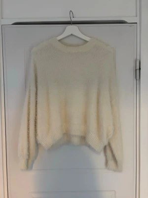 Krämvit fluffig stickad tröja - Mysig oversized stickad tröja i krämvitt med fluffig textur. Tröjan har rund halsringning, långa ärmar och ribbade muddar vid ärmslut och nederkant. Perfekt för lager på lager och en chill look.