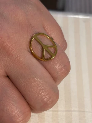 Still l Guldfärgad ring med peace-märke - Cool ring i stål guldfärgad metall med ett stort peace-märke som motiv. Ringen har en öppen design där symbolen är i fokus och ger en retro och statement-känsla. Perfekt för dig som vill sticka ut och visa din personliga stil.