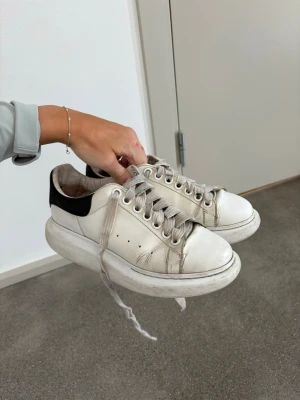 Vita Alexander McQueen sneakers - Chunky vita sneakers från Alexander McQueen med svart detalj på hälen och bred sula. Skorna har gråvita skosnören och perforerade hål på sidan. Klassisk låg modell med rund tå och stilren design.