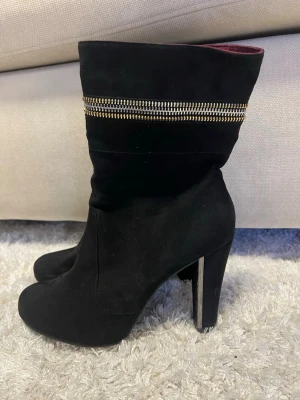 Svarta stövletter med gulddetaljer - Svarta stövletter med höga klackar och dekorativa guldfärgade dragkedjor runt skaftet. Materialet är mocka-liknande och insidan har en mörkröd ton. Stövletterna har en dragkedja på baksidan för enkel på- och avtagning.