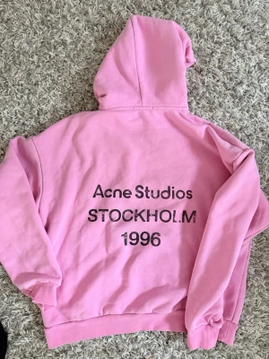 Rosa hoodie från Acne Studios - Säljer en rosa hoodie från Acne Studios med tryck på ryggen där det står 'Acne Studios STOCKHOLM 1996'. Hoodien har huva, ribbade muddar och är gjord i mjukt bomullsmaterial. Perfekt för en chill och trendig look. Har ej bevis på äkthet! Den är unisex 