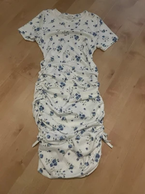 Blommig klänning med snörning - Supersöt klänning i vit med blå blommor och korta ärmar. Tillverkad i mjukt ribbat material som känns bekvämt mot huden.