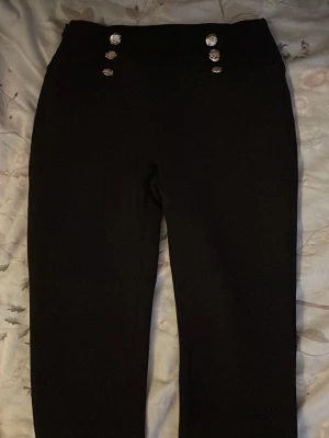 Svarta byxor med dekorativa knappar - Snygga svarta byxor med hög midja och sex dekorativa silverfärgade knappar framtill. Byxorna har en rak passform och är tillverkade i ett mjukt, stretchigt material som ger en bekväm känsla. Perfekta för dig som vill ha en stilren look med en twist.