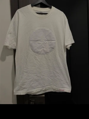 Vit t-shirt från Diamond Supply Co X Travis Scott - Vit t-shirt från Diamond Supply Co med rund hals och ett subtilt cirkelmönster med motiv på bröstet. Liten röd logotypdetalj vid nederkanten. Klassisk passform och tillverkad i mjuk bomull för skön känsla.