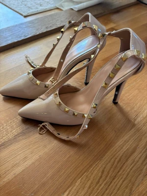 Beige pumps med nitar och remmar - Snygga beige klackar med spetsig tå och höga klackar. Skorna har remmar dekorerade med guldfärgade nitar och spänne runt ankeln. Tillverkade i glansigt material som ger en lyxig känsla. Perfekta för att lyfta din outfit.