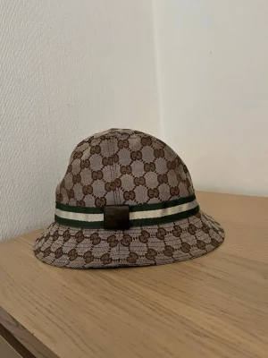 Gucci bucket hat med GG-mönster - Säljer en snygg bucket hat från Gucci i beige och brunt med klassiskt GG-monogram över hela hatten. Hatten har ett grönt och vitt band runtom samt en metallbricka framtill. Tillverkad i canvasmaterial som ger en exklusiv känsla. Den har använts väldigt lite och är i riktigt bra skick!