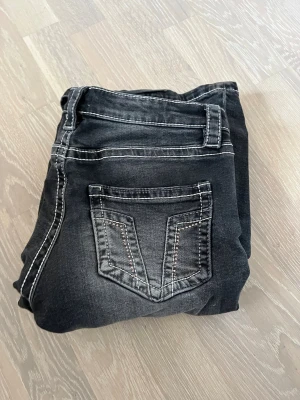 Svarta jeans med kontrastsömmar - Säljer ett par svarta jeans med vita kontrastsömmar och snygga detaljer på bakfickan. Jeansen har klassisk femficksdesign och är tillverkade i denim med en något tvättad look. Perfekta för dig som gillar en stilren men ändå edgy vibe.