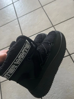 Svarta moon boots med vintertext storlek 38 - Säljer ett par svarta moon boots med fluffig insida och bred sula. Skorna har texten 'WINTER' i vitt runt skaftet och coola detaljer i syntetmaterial. Perfekta för kalla dagar när du vill hålla stilen och värmen.