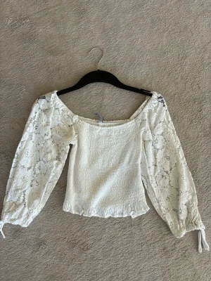 Vit blus med spetsärmar - Söt vit blus med raka axlar och långa, transparenta spetsärmar. Toppen har smockad kropp och volangkant nedtill. Perfekt för dig som gillar romantisk stil och detaljer.