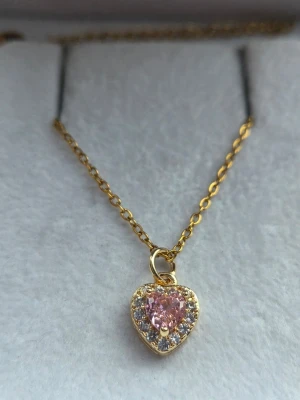 Halsband med hjärthänge och rosa sten - Guldigt stål-halsband med ett hjärtformat hänge. Hänget har en rosa glittrande sten i mitten, omgiven av små klara stenar som ger extra bling. Kedjan är tunn och elegant, perfekt för att lyfta vilken outfit som helst. 