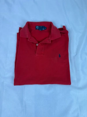 Röd pikétröja från Polo Ralph Lauren - Klassisk röd pikétröja från Polo Ralph Lauren med blå broderad logga på bröstet. Tröjan har krage, knappar framtill och är tillverkad i bomull för en bekväm känsla. Perfekt till jeans eller shorts för en avslappnad stil.