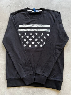 Svart sweatshirt med stjärntryck - L - Cool svart sweatshirt med tryck av vita stjärnor och dollar-mönstrade ränder på bröstet. Tröjan har rund hals, långa ärmar och ribbade muddar vid ärmslut och nederkant. Perfekt för dig som gillar streetwear och grafiska prints.