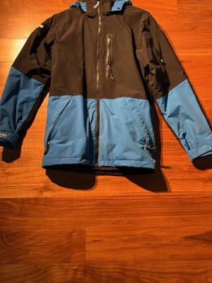 Jacka från 8848 - Snygg vindjacka i blått och svart med dragkedja framtill, huva och flera fickor med dragkedja. Jackan har en sportig look och är tillverkad i ett slitstarkt, vattenavvisande material. Perfekt för blåsiga dagar och utomhusaktiviteter. Dragkedja bröstficka trasig.