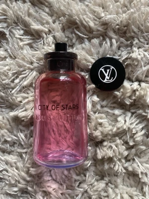 Louis Vuitton City of Stars parfym - Lyxig parfym från Louis Vuitton, City of Stars. Flaskan är i glas med en snygg rosa-lila toning och svart lock med LV-logga. Kommer i en färgglad förpackning med palmmotiv och city lights. Eau de Parfum som känns exklusiv och trendig.