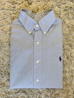 Ljusblå skjorta från Ralph Lauren - Klassisk ljusblå skjorta från Ralph Lauren med diskret rutigt mönster och broderad logga på bröstet. Skjortan har knappar framtill, button-down krage och långa ärmar. Perfekt till jeans eller chinos för en clean och stilren look.