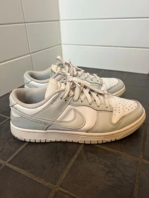 Nike Dunk Low ” White/Photon Dust White” - Nike Dunk Low sneakers i färgen ”White/Photon Dust White”(vit&grå) med klassiska nike märket på sidan. Lågt skaft och vita skosnören. Stilren och tidlös modell som passar till många olika outfits, både för herr och dam😃