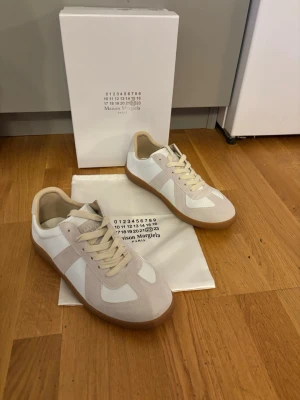 Maison Margiela vita sneakers - Snygga vita sneakers från Maison Margiela med beige mockadetaljer och klassisk gummisula. Skorna har en låg silhuett, vita skosnören och stilren design som passar till det mesta. Materialmix av läder och mocka ger en exklusiv känsla.