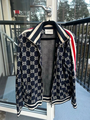 Mörkblå GG-mönstrad track jacket Gucci - Snygg mörkblå track jacket från Gucci med vitt GG-mönster över hela jackan. Jackan har vita och röda ränder på ärmarna, ribbade muddar och krage med breda ränder samt dragkedja framtill. Perfekt för dig som vill sticka ut med en ikonisk streetwear-look. 